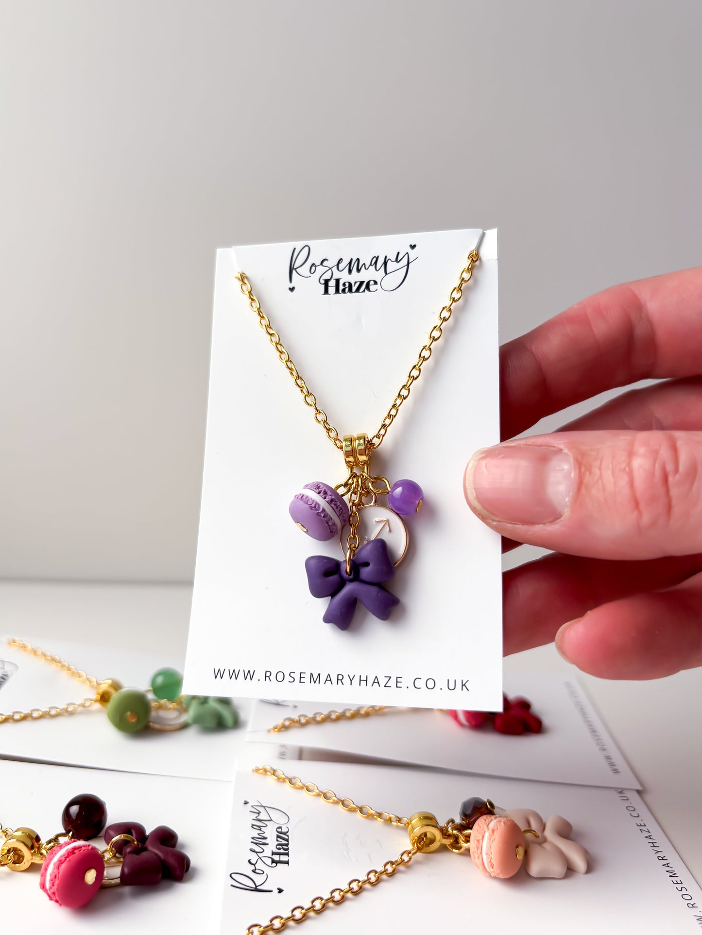 A hand holds a gold necklace on a white display card featuring a purple macaron and bow charm. The card is branded Rosemary Haze, and other colorful necklace charms are visible on the table below against a light gray background.
